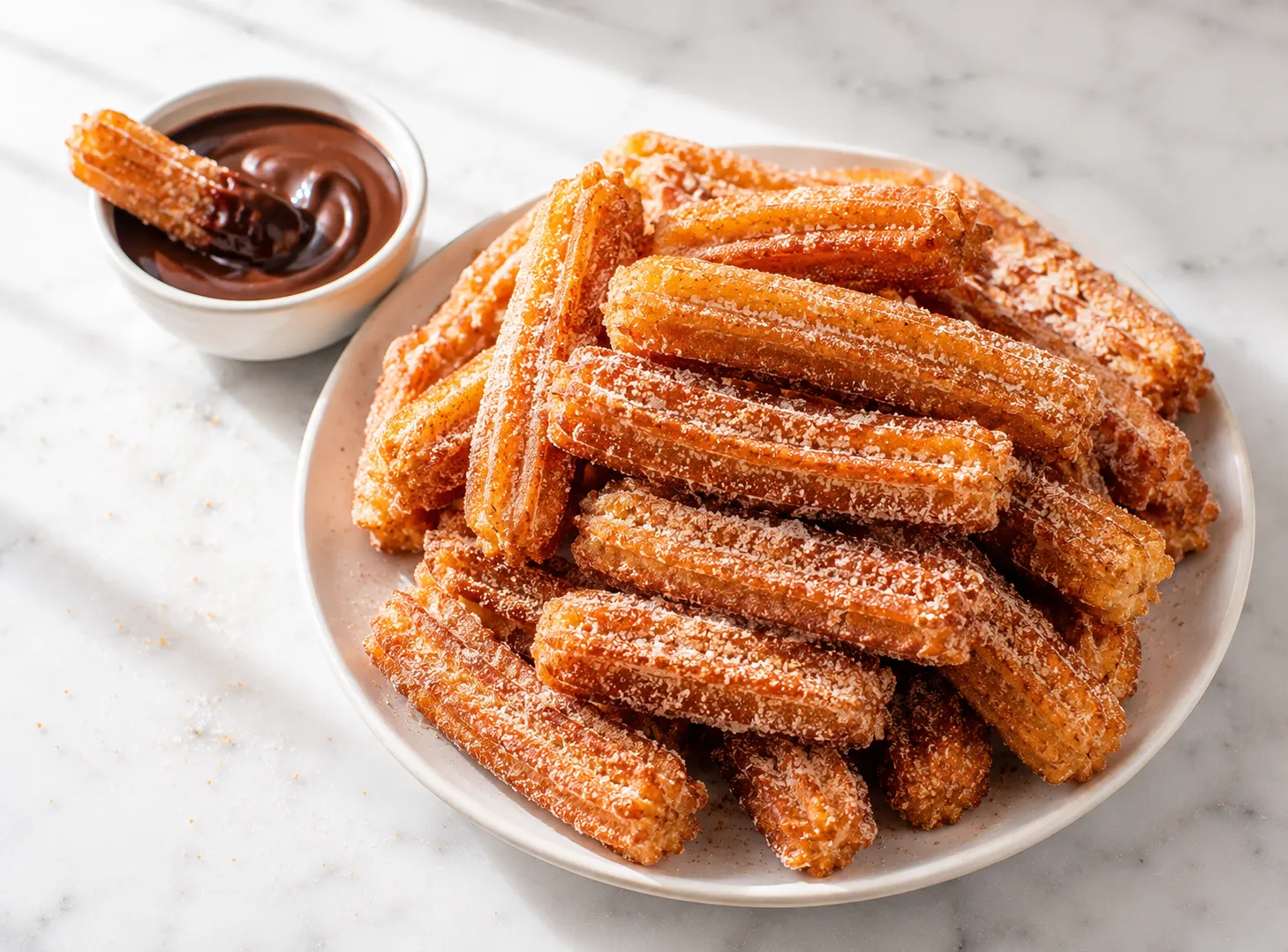 Crispy cinnamon churros piled on a white plate with a bowl of chocolate dipping sauce on a white marble surface in natural daylight