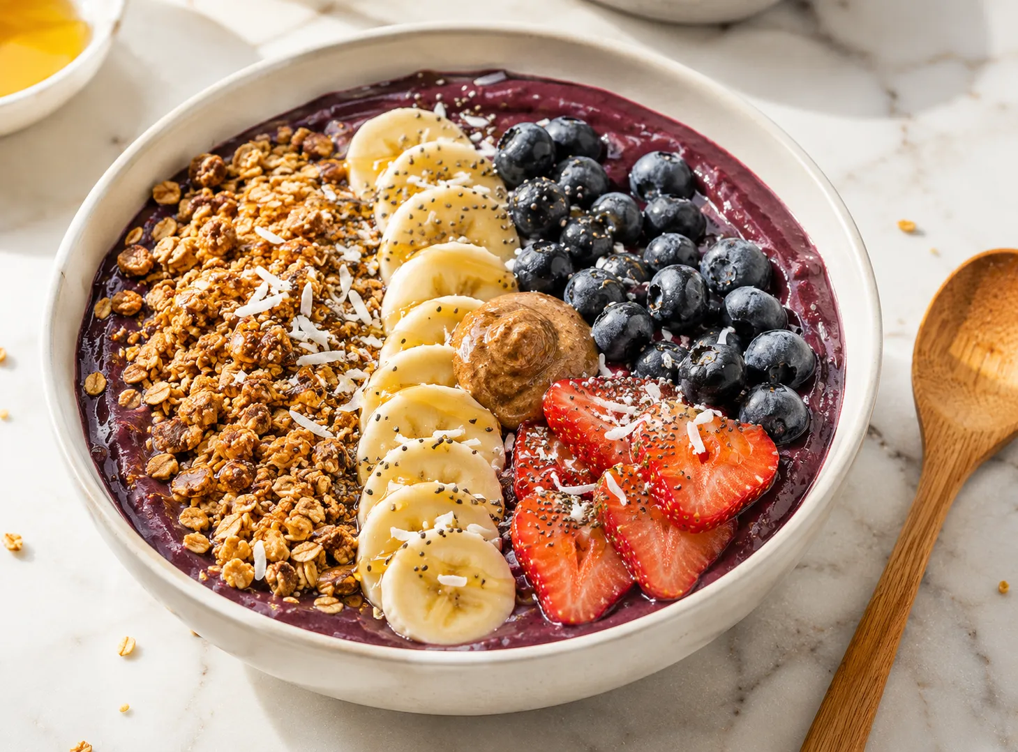 Easy acai bowl recipe with thick purple acai base topped with fresh banana, blueberries, strawberries, granola, chia seeds, and almond butter in a white ceramic bowl on white marble surface