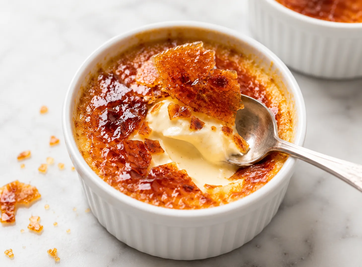 Overhead view of a cracked creme brulee with a spoon lifting the caramel sugar crust showing the silky pale custard underneath on a white marble surface.