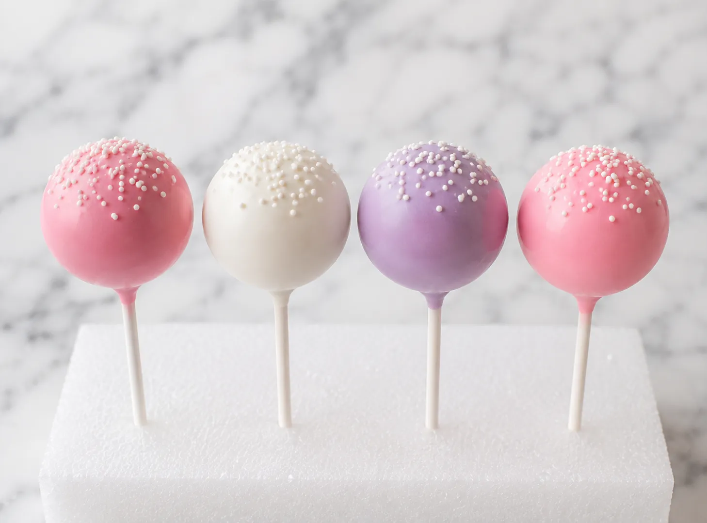Twenty-four bite-sized cake pops with smooth pink, white, and lavender candy melt coating standing upright in a white block on a marble surface in soft natural light