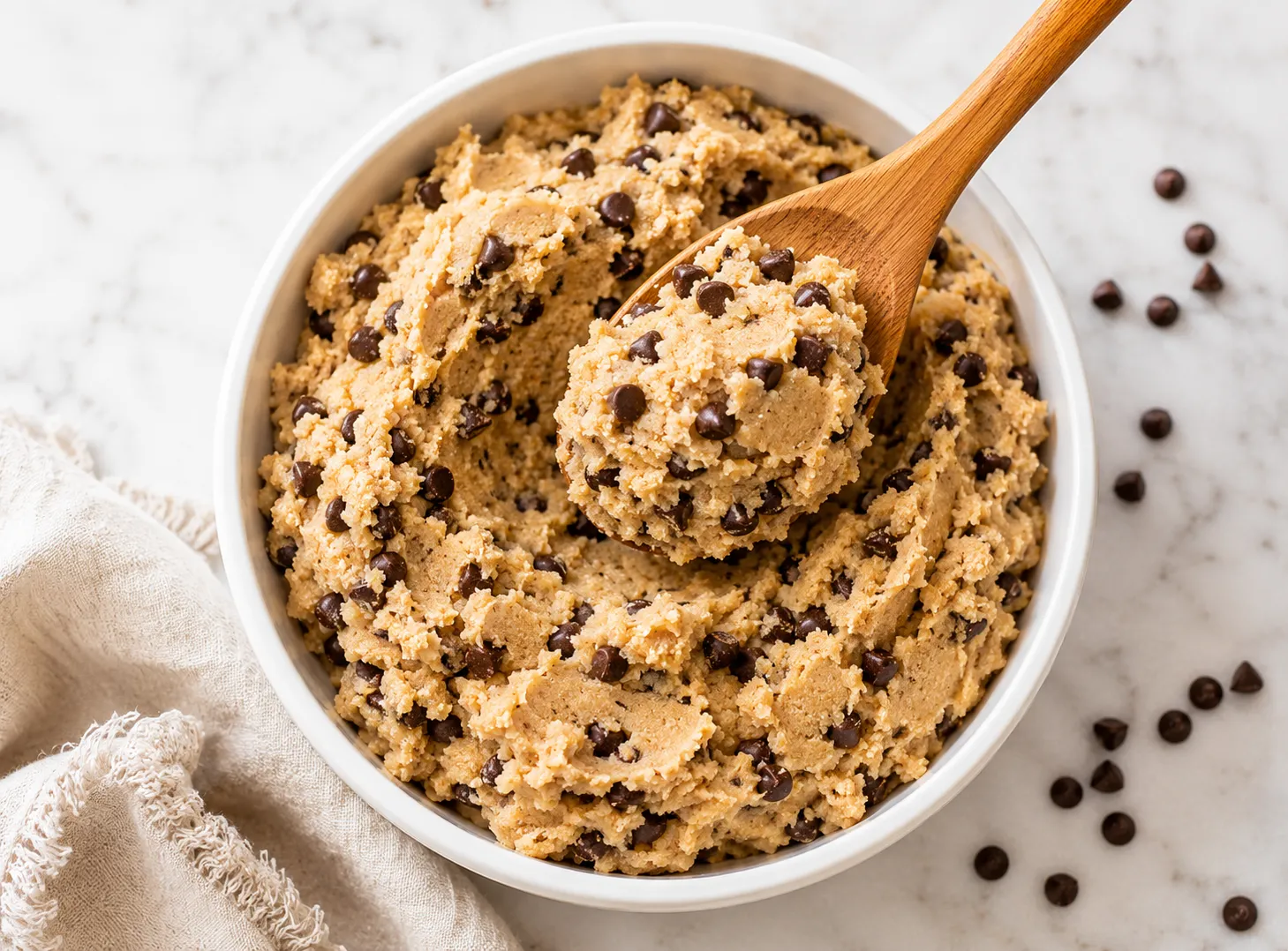 White ceramic bowl of edible cookie dough loaded with mini chocolate chips on white marble surface with wooden spoon